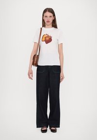 Witte t-shirt met een grote aardbeienafbeelding, gecombineerd met losse donkere denim jeans met groene stiksels en een bruine schoudertas.