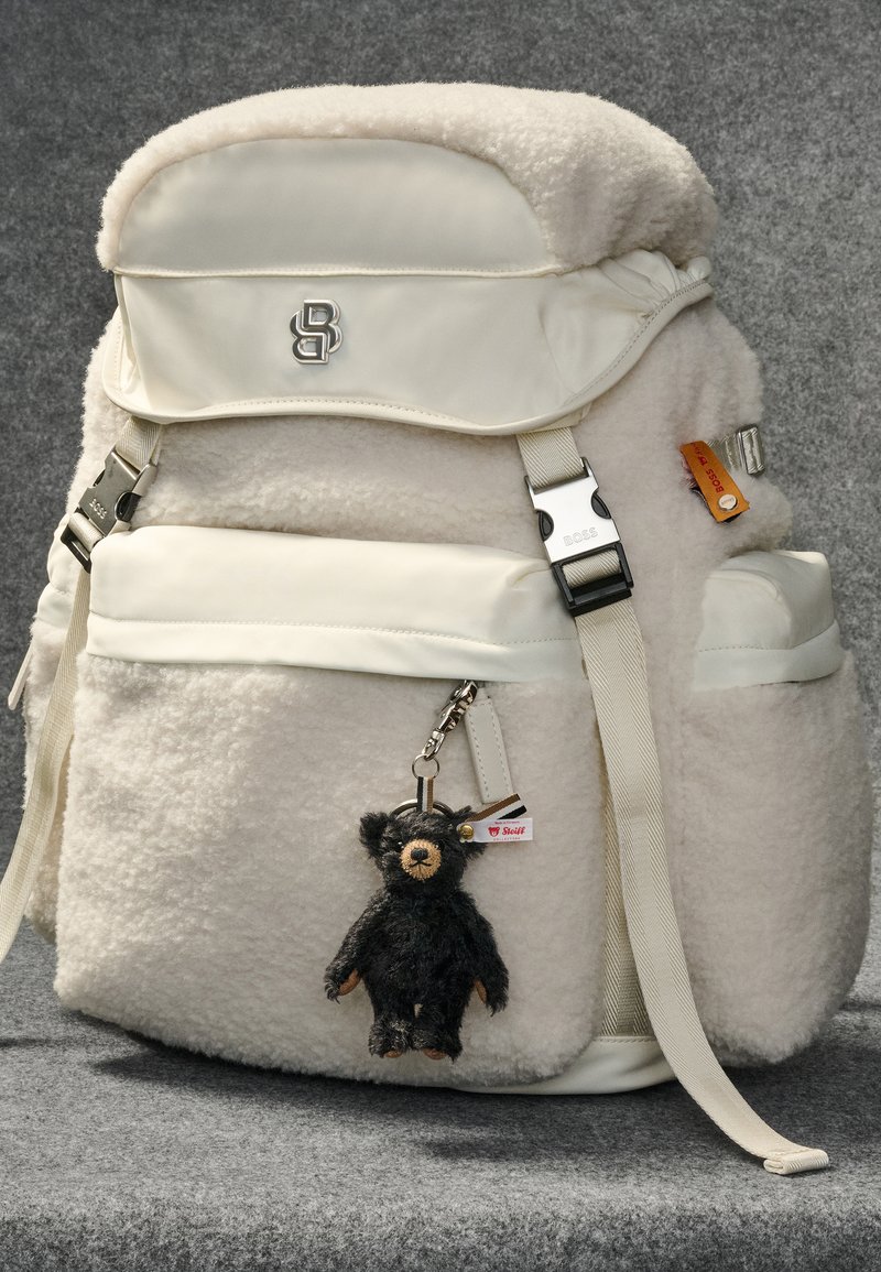 Mochila de felpa blanca con parte superior lisa, correas ajustables, herrajes plateados y un llavero de oso negro adjunto como detalle.