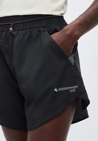 Κοντινό πλάνο ενός ατόμου που φοράει μαύρα σορτς υπαίθρου με ελαστική μέση και το χέρι στην πλαϊνή τσέπη, με την επωνυμία "Blåhammarus Diser" στο πόδι.