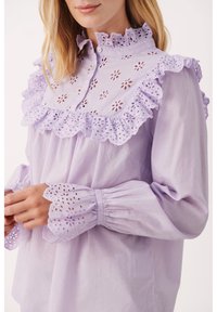 Blouse lavande avec un col à volants et des découpes florales, manches longues avec poignets élastiques et délicats détails ajourés sur les bords.