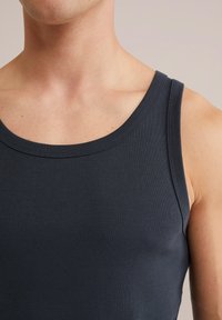 Torso masculino con una camiseta sin mangas de color azul marino oscuro, mostrando el cuello, el hombro y la parte superior del pecho contra un fondo neutro.