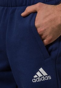 Käsi taskus tumesinistes Adidas spordipükstes, logo reie peal, nähtav elastne vöökoht.