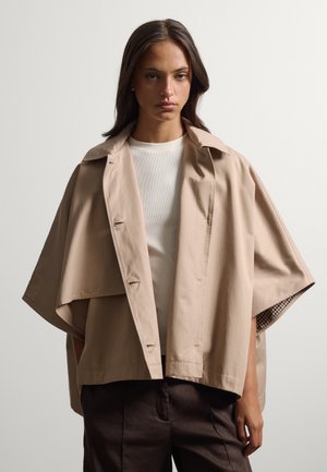 Frau trägt einen beigen Trenchcoat mit kurzen Ärmeln über einem weißen Top und dunklen Hosen vor einem einfarbigen, hellen Hintergrund.