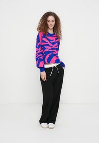 Φούξια και μπλε πουλόβερ με μοτίβο ρίγες ζέβρας, μακριά μανίκια και χαλαρή εφαρμογή, συνδυασμένο με φαρδιά μαύρη παντελόνια και λευκά αθλητικά παπούτσια.