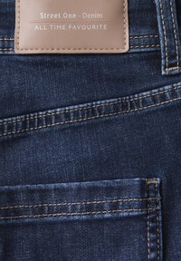 Tissu en denim bleu foncé avec une poche arrière, des détails cousus et une étiquette en cuir beige avec "Street One - Denim" et "ALL TIME FAVOURITE."