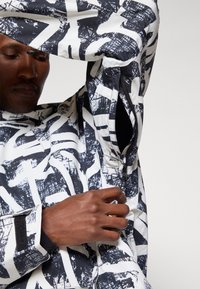 Μαύρο και άσπρο μ patterned σακάκι με τολμηρά αφηρημένα σχέδια, μακριά μανίκια, φερμουάρ και λεία, γυαλιστερή επιφάνεια.