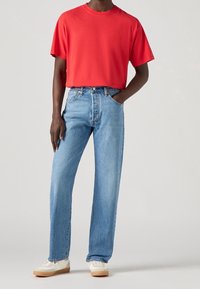 Uomo che indossa una t-shirt rossa a maniche corte, jeans dritti di colore blu chiaro e tennis bianchi con suola in gomma, in piedi con una mano in tasca.
