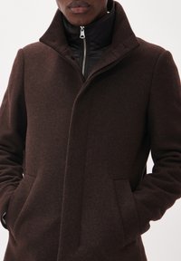 Bruine wollen jas met een hoge kraag en een gevoerde zwarte binnenlaag met rits, voorzien van zijzakken en een textuurrijke stofafwerking.