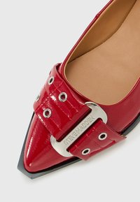 Chaussure plate pointue en cuir verni rouge avec une grande boucle comportant des œillets en argent et une plaque gravée "Steve Madden".