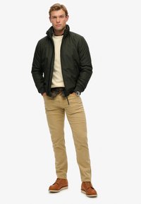 Dunkelgrüne Steppjacke, cremefarbener Pullover, darunter ein kariertes Hemd, beige Chinos und braune Schnürschuhe. Modell steht mit den Händen in den Taschen.