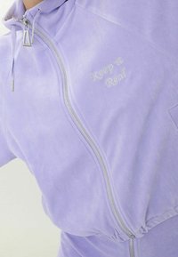 Lila zip-jacka med silverdetaljer, med broderad text "Keep it Real". Mjuk textur och gathered midjedetalj.