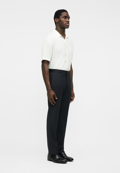 Homme debout, portant une chemise blanche à manches courtes à boutons, un pantalon noir cintré et des chaussures habillées noires, sur un fond clair uni.