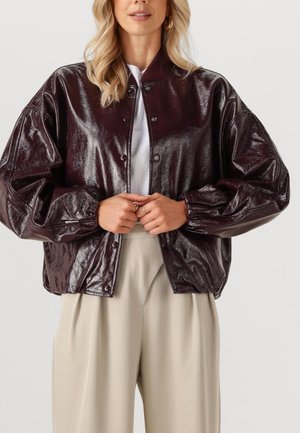 Burgunderfarbene lackierte Bomberjacke mit einer Knopfverschlussfront, übergroßen Ärmeln und gerippten Bündchen. Getragen über einem weißen Oberteil und beigen Hosen.