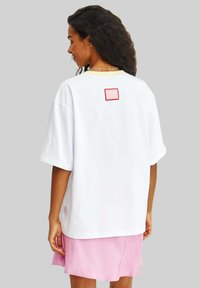 Bílé oversized tričko s krátkými rukávy, na zadní straně s červeným obdélníkovým nášivkem. Nosené s světle růžovou sukní.