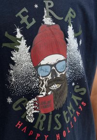 Un squelette avec une barbe, un bonnet rouge et des lunettes de soleil tenant une tasse fumante avec écrit "mettons-nous à l'aise", avec des arbres enneigés et un texte festif.