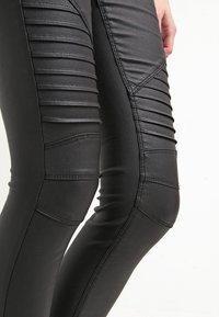 Las leggings sintéticas negras presentan paneles horizontales acolchados a lo largo de los muslos, con una textura brillante y costuras estructuradas.