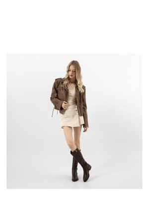 Jeune femme portant une veste en cuir marron, une jupe beige et des bottes montantes marron foncé, debout les jambes croisées sur un fond blanc.