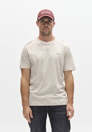 Muž v béžovom tričku s logom „Gabba“, tmavých džínsoch a bordovej šiltovke „Noosa Beach“ stojí na bielom pozadí.