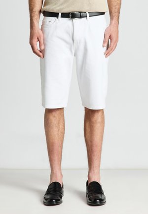 Uomo che indossa pantaloncini bianchi fino al ginocchio, cintura nera, top beige in maglia e scarpe slip-on in pelle nera, in piedi davanti a uno sfondo semplice.