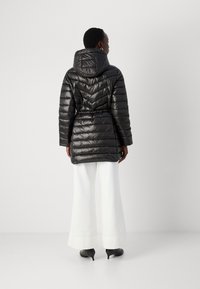 Doudoune noire avec capuche, dotée d'un motif matelassé, d'une taille ajustable avec cordon de serrage et d'une texture lisse, assortie à un pantalon large blanc et des talons.