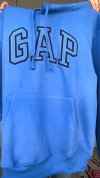 Modrá mikina s kapucí s přední kapsou a velkým našitým logem GAP v světle modré barvě s černým obrysem, držená za ramena rukama.