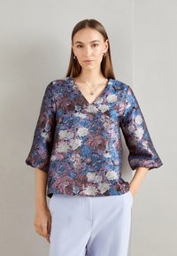 Blouse florale en tissu texturé avec un décolleté en V, des manches trois-quarts, et des couleurs violet, bleu et blanc sur un fond sombre.