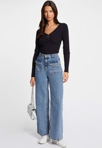 Schwarzes Langarmoberteil mit strukturiertem Design, kombiniert mit hochgeschnittenen, weit geschnittenen blauen Jeans mit Fronttaschen und ausgefranstem Saum. Silberne Kettenhandtasche.