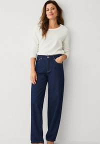Jeans a vita alta e gamba larga in denim blu scuro con un classico design a cinque tasche e dettagli di cucitura sottili. Abbinati a un maglione crema.