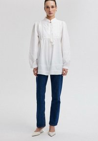 Blusa bianca di cotone con un collo alto, dettagli plissettati e maniche lunghe e voluminose. Abbinata a jeans blu dritti e tacchi a punta.