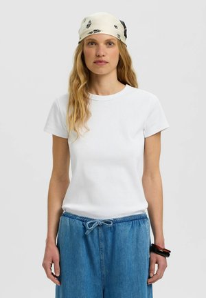 Kobieta o długich blond włosach, ubrana w kremową bandanę, białą koszulę z krótkim rękawem, niebieską jeansową spódnicę ze sznurkiem oraz czarne i brązowe bransoletki.