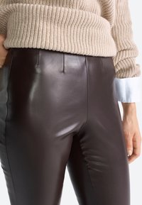 Pantaloni in pelle marrone con una finitura lucida, vita alta e cuciture minime, abbinati a un maglione beige texture e dettagli azzurri sulle maniche.