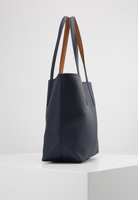 Sac en toile bleu marine avec surface texturée et doubles poignées marron, posé sur un piédestal blanc contre un fond neutre.