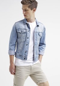 Hellblaue Jeansjacke mit Knopfverschluss, zwei Brusttaschen und hochgekrempelten Ärmeln, kombiniert mit einem weißen Hemd und beigen Shorts.