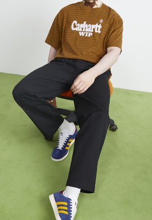 Camiseta marrón a rayas con logo blanco, pantalones negros, zapatillas azules con rayas amarillas y detalles blancos, sentado en una silla naranja.