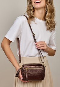 Smeđi metalik crossbody torba s dva zipovana odjeljka, zlatnim detaljima i naglaskom s resicama. Podesiva naramenica, pravokutnog oblika.