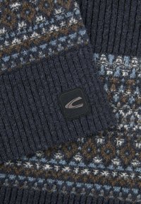 Nærbillede af en mørkeblå uldtrøjeslange med ribbet manchet og geometrisk mønster i blå, hvid og brun farver, med et sort logo-patch.