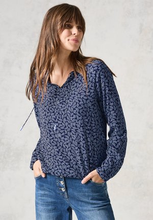 Blusa blu navy con un motivo astratto di foglie bianche, caratterizzata da un scollo a V, maniche lunghe e una vestibilità rilassata. Indossata con jeans blu.