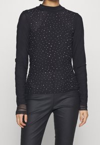 Haut noir à manches longues avec un design ajusté, comportant des clous irisés dispersés et des accents en maille transparente le long des manches.