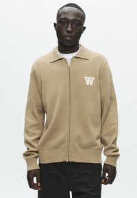 Pull beige à fermeture éclair avec col et ourlet côtelé, arborant un logo blanc en forme de "W" sur le côté gauche de la poitrine. Tissu lisse avec manches longues.