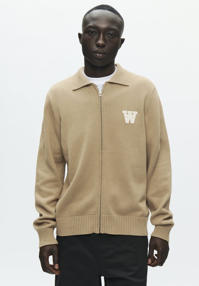 Pull beige à fermeture éclair avec col et ourlet côtelé, arborant un logo blanc en forme de "W" sur le côté gauche de la poitrine. Tissu lisse avec manches longues.