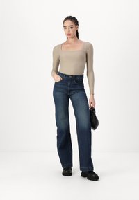 Beige Langarmbodysuit, hochgeschnittene dunkelblaue Jeans, schwarze Handtasche und schwarze Schuhe. Das Model steht zentriert vor einem weißen Hintergrund.
