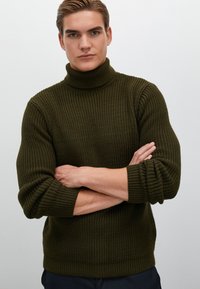 Vihreä oliivinvärinen poolopaita, jossa on tekstuuri neulekuvio, pitkät hihat sekä ribbitut hihat ja helma, henkilö, jolla on kädet ristissä.