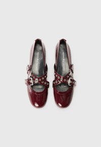 Scarpe in pelle verniciata bordeaux con punta rotonda e cinturini alla caviglia, dotate di hardware argentato e ritagli circolari. Texture liscia e lucida.