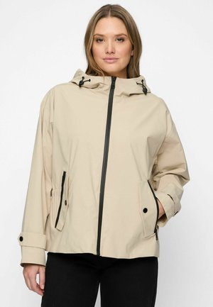 Femme portant une veste beige à capuche avec une fermeture éclair et des boutons noirs, mains dans les poches, sur un fond blanc uni.