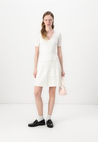 White knit dress with short sleeves, textured patterns, and a V-neckline. Paired with black shoes, light gray socks, and a small pink handbag.