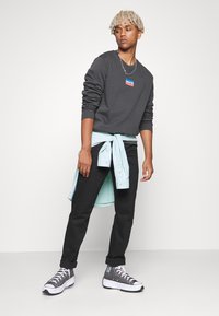 Sudadera gris con el logo de Levi's, pantalones negros, camisa ligera azul claro en capas atada a la cintura, zapatillas grises y blancas, fondo neutro.