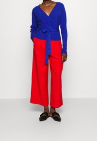 Person in einem blauen Wickel-Pullover mit einem Bindegürtel, hellroten weit geschnittenen Hosen und schwarzen Slippern, die vor einem schlichten Hintergrund steht.