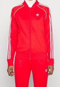 Giacca della tuta Adidas rossa con zip frontale, strisce bianche lungo spalle e maniche, polsini a costine e logo sul petto.