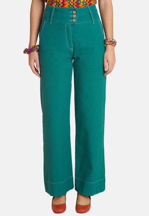 Hochtaillierte, weit geschnittene Hose in Türkis mit drei Knöpfen vorne, kombiniert mit bunten Armbändern und roten Schuhen.