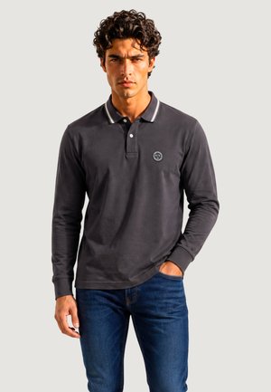 Uomo con i capelli ricci che indossa una polo scura a maniche lunghe e jeans blu, in piedi con una mano in tasca davanti a uno sfondo semplice.